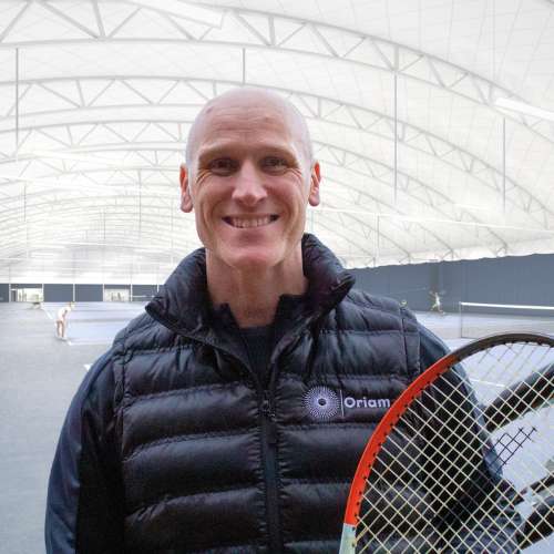 Picture of Danny Curtis holding a tennis racket with an indoor tennis court in the background
