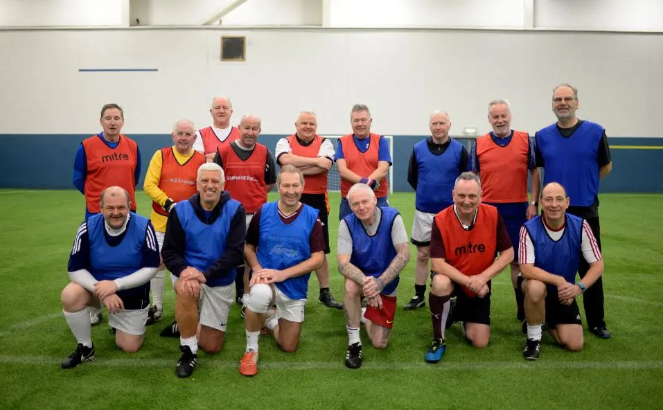 Walking Football Team line-up photo. Two rows of football player in read and blue bibs. Front row kneeling, Back row standing. green synthetic pitch in background. 