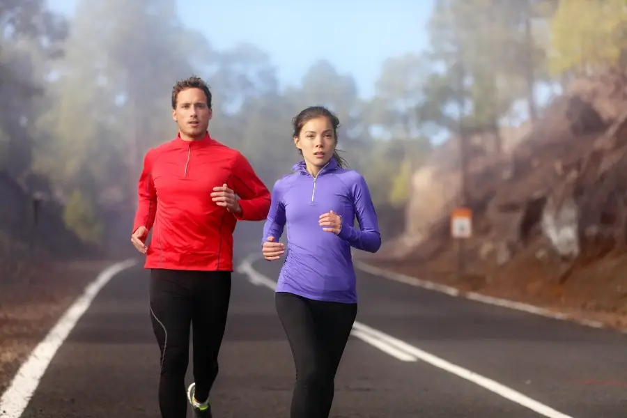 Two people running outdoors in autumn scenery. Runner 1 is small female, brown hair, purple top. Runner 2 is taller male, wearing red top.