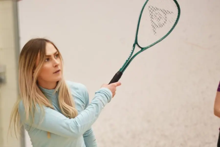 link opens in new tab. Close-up photo of Female playing squash. Blonde Hair, light turquoise top, green squash racket at the top of a backhand swing.