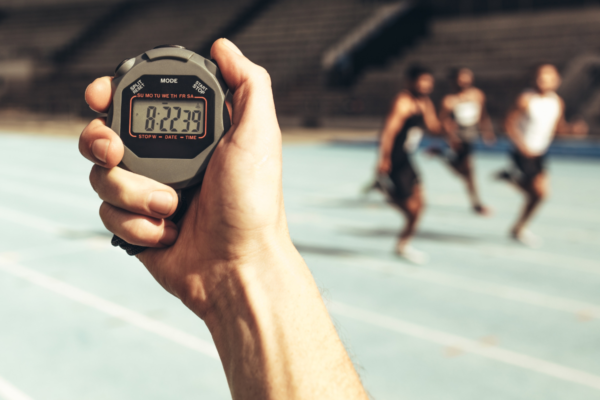 Foreground. Sharp Focus Close-up of person's hand holding  black and silver stopwatch displaying 8.22. Background: Soft focus blur, 3 runners on running track. 