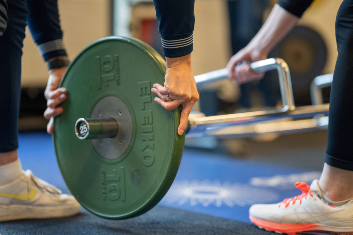 Close-up image of person loading a green 10kg weight onto a bar in Oriam S&C Gym with another person supporting. 