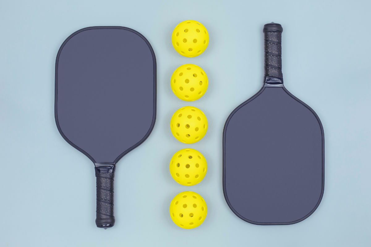 Play Pickelball at Oriam. Light blue background with two grey pickleball rackets and a row of yellow pickleballs between them.