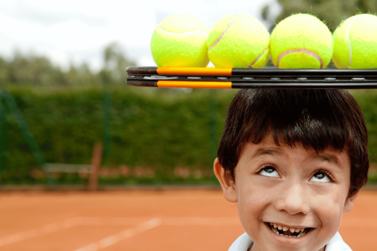 Foreground. Boy on righthand side of image looking up as he balances a racket full of tennis balls on his head. Background red clay tennis court with green hedges. 
