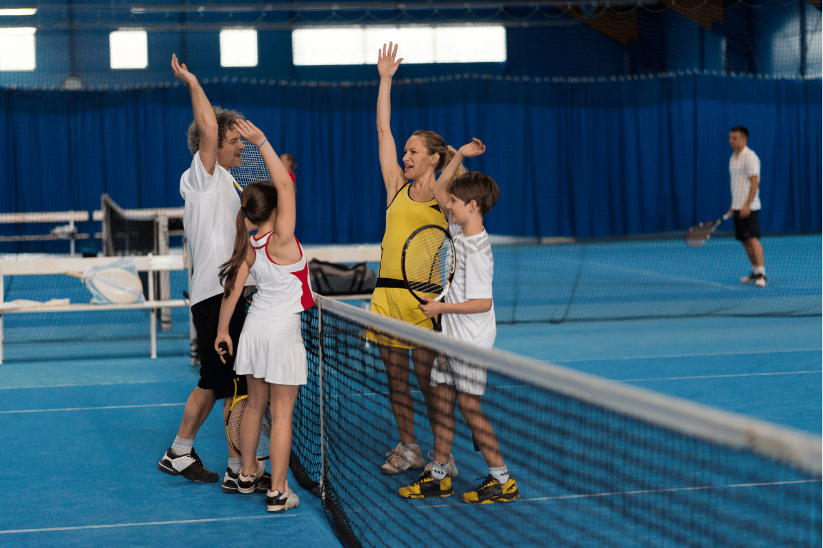 Blue indoor tennis court with blue curtain backdrop. Four people on court, celebrating end of tennis match, left side of net adult male and junior female. Right side of net adult female and junior male. all players facing each other giving high fives, representing good sportsmanship
