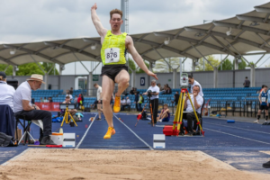 Action shot of Long jump Athlete mid-jump into sandpit. Male athlete wearing yellow vest, black shorts and orange trainers. background stadium with blue track and blue seats.