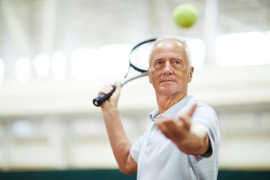 Male Tennis Player facing camera. White hair, white polo-shirt. Serving a tennis ball overhand. 