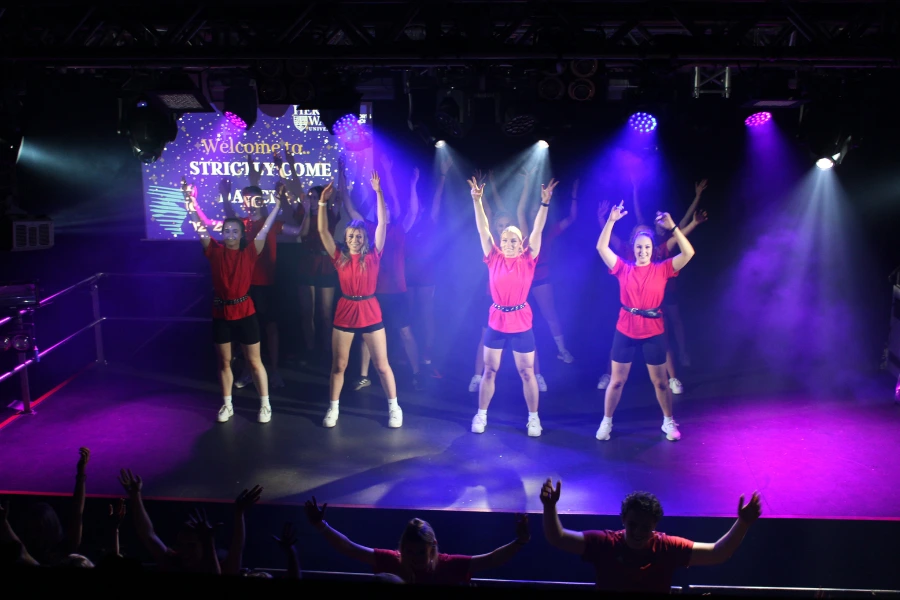 4 dancers wearing red tops and black shorts on stage. blue and pink lighting. 