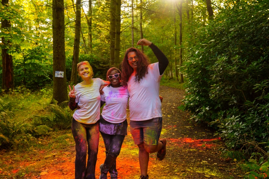 Three student stood posing on the running trail at HWU Edinburgh Campus. All wearing white t-shirts covered in colourful powder.